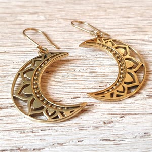 Può includere: Un paio di orecchini a mezzaluna color oro. Ogni orecchino presenta un elaborato motivo floreale all'interno della forma della luna e un bordo di perline. Gli orecchini hanno una chiusura a gancio. Su una superficie di legno chiaro.