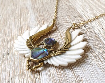 May include: A gold necklace with a white and gold pendant featuring a scarab beetle with iridescent abalone shell accents.