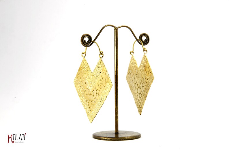 May include: A pair of gold-tone earrings with a diamond shape and intricate cut-out design. The earrings are hanging from a stand.