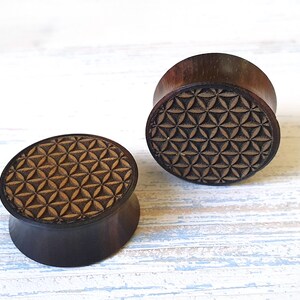 May include: Two wooden ear gauges with a geometric pattern carved into the top. The pattern is a flower of life design.