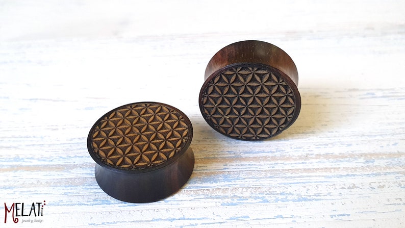 May include: Two wooden ear gauges with a geometric pattern carved into the top. The pattern is a flower of life design.