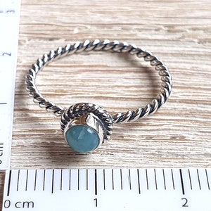 May include: A silver ring with a twisted band and a light blue gemstone set in a silver bezel. The ring is measured against a ruler in centimeters.