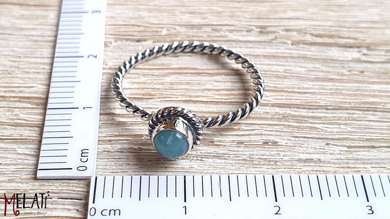 May include: A silver ring with a twisted band and a light blue gemstone set in a silver bezel. The ring is measured against a ruler in centimeters.