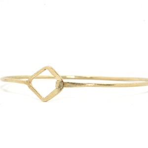 May include: A gold-coloured bangle bracelet featuring a square-shaped central design. The bracelet has a simple, elegant design with a textured finish. The jewellery is displayed on a white background.