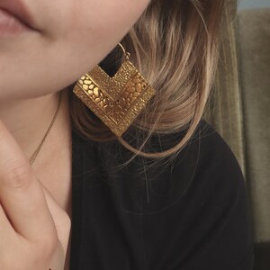 May include: Gold geometric earrings with a textured surface. The earrings are in the shape of a square with a pointed bottom.