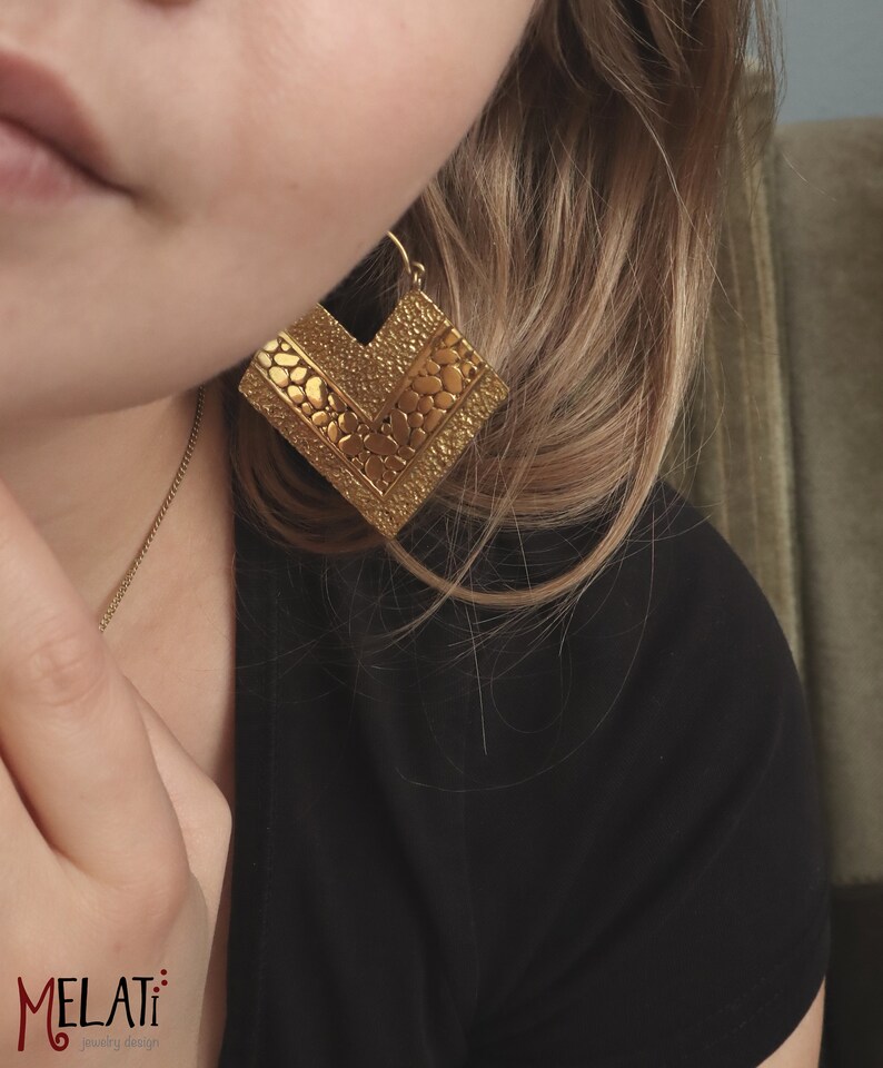 May include: Gold geometric earrings with a textured surface. The earrings are in the shape of a square with a pointed bottom.
