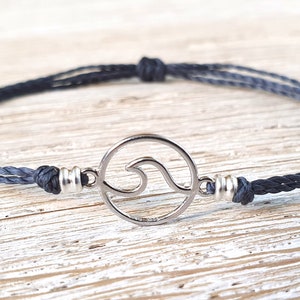 May include: A dark gray braided cord bracelet with a silver wave charm. The charm is a circle with a cut-out wave design.