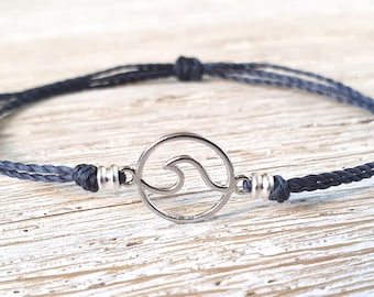 May include: A dark gray braided cord bracelet with a silver wave charm. The charm is a circle with a cut-out wave design.