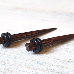 May include: A pair of dark brown, tapered ear stretching tapers with a smooth, polished finish. Each piece features a single black rubber O-ring positioned near the thicker end. The text MELATI jewelry designer is visible in the bottom left corner.
