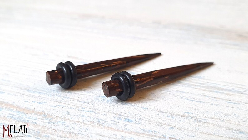 May include: A pair of dark brown, tapered ear stretching tapers with a smooth, polished finish. Each piece features a single black rubber O-ring positioned near the thicker end. The text MELATI jewelry designer is visible in the bottom left corner.