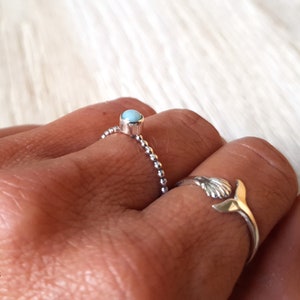 May include: Two silver rings are displayed on a hand. One ring features a small, round, light blue stone. The other ring has a seashell design with a gold-colored tail. The rings are on a light-colored surface.