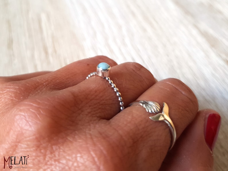 May include: Two silver rings are displayed on a hand. One ring features a small, round, light blue stone. The other ring has a seashell design with a gold-colored tail. The rings are on a light-colored surface.