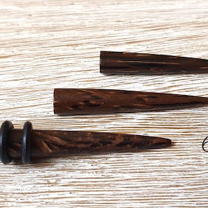 May include: Three dark brown wooden ear tapers with a natural wood grain pattern. The bottom taper features two black rubber O-rings. The text 6 mm is printed on the right side, and the word MELATI is printed in the bottom left corner.