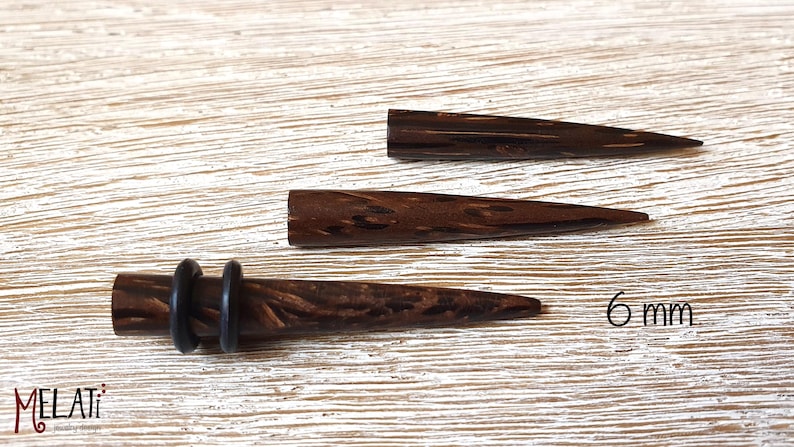 May include: Three dark brown wooden ear tapers with a natural wood grain pattern. The bottom taper features two black rubber O-rings. The text 6 mm is printed on the right side, and the word MELATI is printed in the bottom left corner.