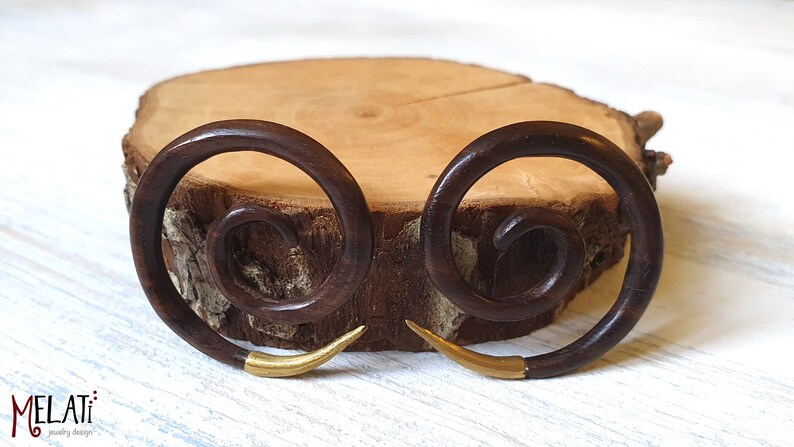 May include: A pair of brown wooden spiral earrings with gold accents. The earrings are displayed on a wooden surface.