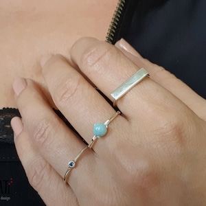 Puede incluir: Tres anillos de plata en un dedo. El anillo superior es una banda plana y rectangular. El anillo del medio presenta una piedra preciosa azul claro. El anillo inferior tiene una pequeña piedra preciosa azul oscuro.