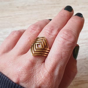 Könnte beinhalten: Ein goldfarbener Ring mit einem geometrischen Design aus geschichteten Quadraten und Rechtecken. Der Ring wird an einer Hand mit dunklem Nagellack getragen. Das Design des Rings ist der Mittelpunkt.