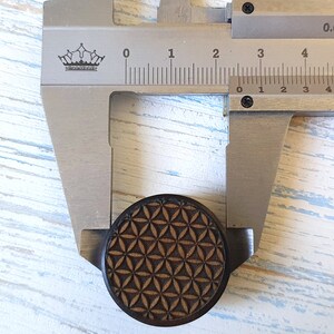 May include: A close-up of a wooden ear gauge with a geometric pattern, measured with calipers to be 2 cm in diameter.