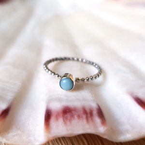 May include: A silver ring with a beaded band and a light blue, oval-shaped gemstone. The ring is set against a white background, possibly a seashell. The ring's design is simple and elegant.