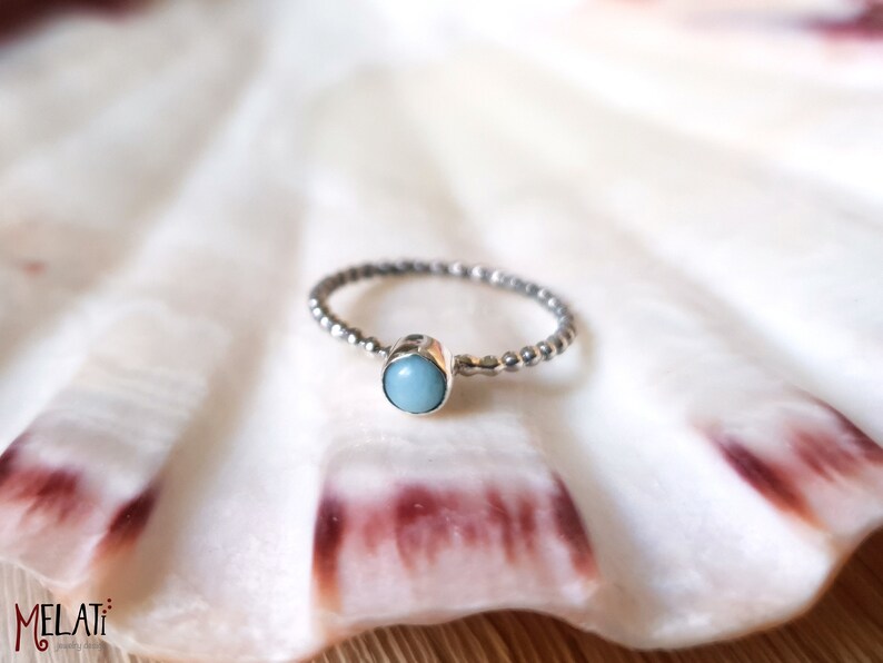 May include: A silver ring with a beaded band and a light blue, oval-shaped gemstone. The ring is set against a white background, possibly a seashell. The ring's design is simple and elegant.
