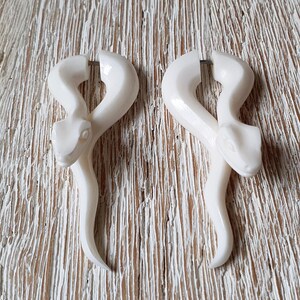 Può includere: Due orecchini color avorio a forma di serpente. Ogni orecchino presenta una testa di serpente dettagliata e un corpo curvo. Gli orecchini sono esposti su una superficie chiara e strutturata. Un accessorio unico.