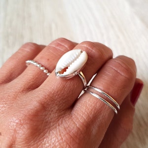 Könnte beinhalten: Ein silberner Ring mit einem weißen Kaurischalen-Design. Der Ring wird an einem Finger mit anderen silbernen Ringen getragen.