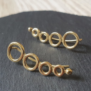 Op de afbeelding: Twee gouden oormanchetten met een gebogen ontwerp met meerdere verbonden cirkels. De oormanchetten liggen op een zwart oppervlak.