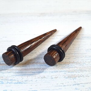 May include: A pair of dark brown wood ear tapers with a polished finish and a pointed tip. Each taper features a flat circular base with a black rubber O-ring secured around the circumference. The text MELATI jewelry design is visible in the corner.