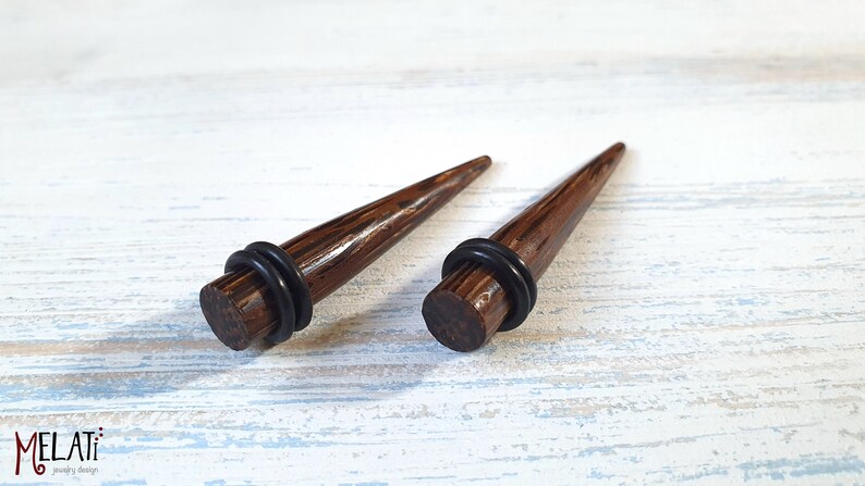 May include: A pair of dark brown wood ear tapers with a polished finish and a pointed tip. Each taper features a flat circular base with a black rubber O-ring secured around the circumference. The text MELATI jewelry design is visible in the corner.