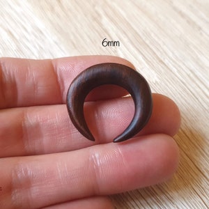 May include: A 6mm crescent moon shaped wooden ear gauge, shown in a hand. The gauge is a dark brown color.