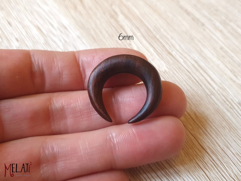 May include: A 6mm crescent moon shaped wooden ear gauge, shown in a hand. The gauge is a dark brown color.