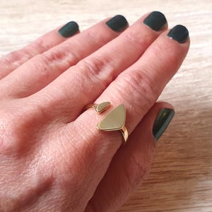 Könnte beinhalten: Ein goldfarbener Ring mit geometrischem Design wird an einem Finger getragen. Der Ring hat eine kleine, abgerundete Form und eine größere, dreieckige Form. Die Hand der Person hat dunkelgrünen Nagellack. Der Ring ist ein Schmuckstück.