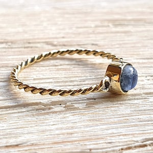 Puede incluir: Un anillo de oro con una banda trenzada y una piedra preciosa azul. El anillo está sobre una superficie de madera clara.
