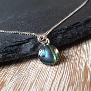Könnte beinhalten: Eine silberne Halskette mit einem runden, schillernden Abalone-Muschel-Anhänger. Der Anhänger zeigt Farbtöne von Blau, Grün und Gold. Die Halskette wird auf einer dunklen, strukturierten Oberfläche präsentiert.