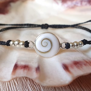 Può includere: Un braccialetto in cordoncino nero con un ciondolo a conchiglia argentato e bianco con un motivo a spirale. Il braccialetto è decorato con perline argentate.