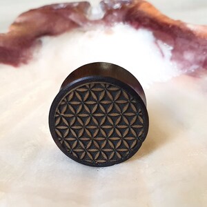 May include: A dark brown wooden ear plug with a geometric flower of life design carved into the surface.