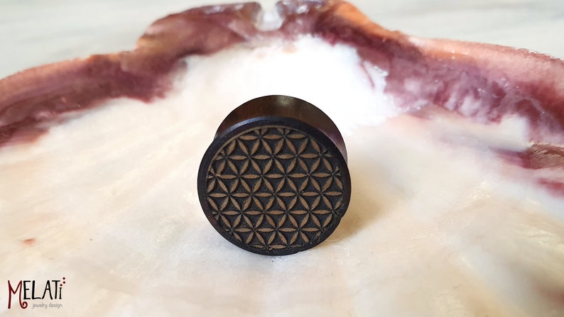 May include: A dark brown wooden ear plug with a geometric flower of life design carved into the surface.