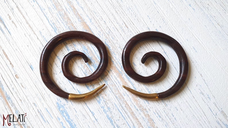 May include: A pair of dark brown wooden spiral ear gauges with gold metal accents. The gauges are on a white wooden surface.
