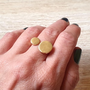 Könnte beinhalten: Ein goldfarbener Ring mit zwei kreisförmigen Elementen, einem kleinen und einem großen, wird an einem Finger getragen. Der Ring ist ein minimalistisches Design. Die Hand hat dunklen Nagellack. Schmuck von Melati jewelry design.