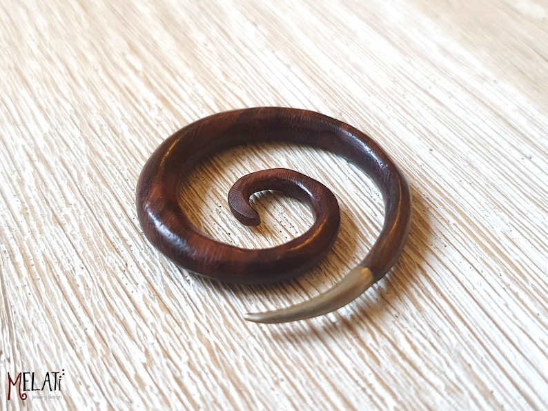 May include: A dark brown wooden spiral ear gauge with a silver metal spike. The gauge is made of wood and has a spiral design. The spike is made of metal and is attached to the end of the gauge.