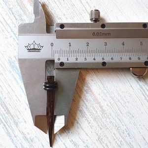 May include: A dark brown, tapered wooden ear plug with two black rubber O-rings, positioned within a metal vernier caliper. The caliper displays a measurement of approximately 0.3 inches, indicating the diameter of the jewelry piece.