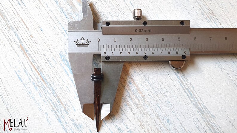 May include: A dark brown, tapered wooden ear plug with two black rubber O-rings, positioned within a metal vernier caliper. The caliper displays a measurement of approximately 0.3 inches, indicating the diameter of the jewelry piece.
