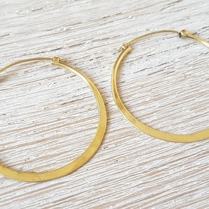 Könnte beinhalten: Ein Paar großer, goldfarbener Creolen. Die Ohrringe haben ein flaches, breites Design und einen einfachen Verschluss. Sie werden auf einer hellen, strukturierten Oberfläche präsentiert.