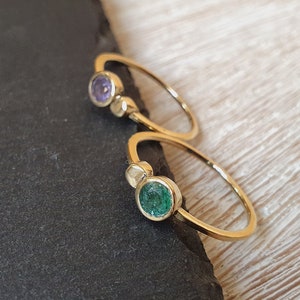 Puede incluir: Dos anillos de oro con pequeñas piedras redondas. El anillo superior tiene una piedra morada y el anillo inferior tiene una piedra verde.
