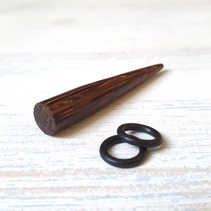 May include: A dark brown wooden taper with a visible wood grain pattern and two small black rubber O-rings. The text MELATI jewelry design is visible in the bottom left corner of the image, featuring a stylized red letter M and black text.