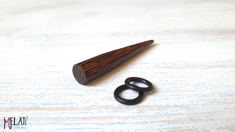 May include: A dark brown wooden taper with a visible wood grain pattern and two small black rubber O-rings. The text MELATI jewelry design is visible in the bottom left corner of the image, featuring a stylized red letter M and black text.