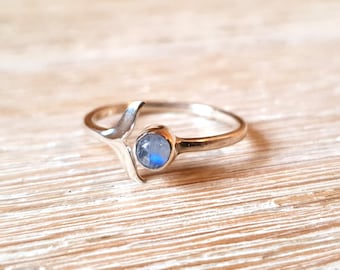 Può includere: Un anello d'argento con una pietra di luna blu incastonata in una fascia semplice. L'anello ha un design unico con una linea curva che avvolge la pietra.