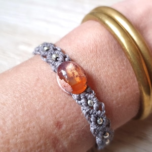 Può includere: Un braccialetto in macramé grigio con un accento di perline argentate e una pietra preziosa ovale arancione e marrone. Il braccialetto è indossato su un polso con un braccialetto d'oro.