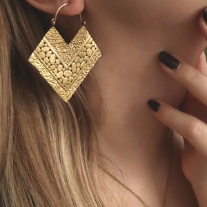 May include: Gold statement earrings with a geometric, textured design. The earrings are shaped like a chevron and have a hammered finish.