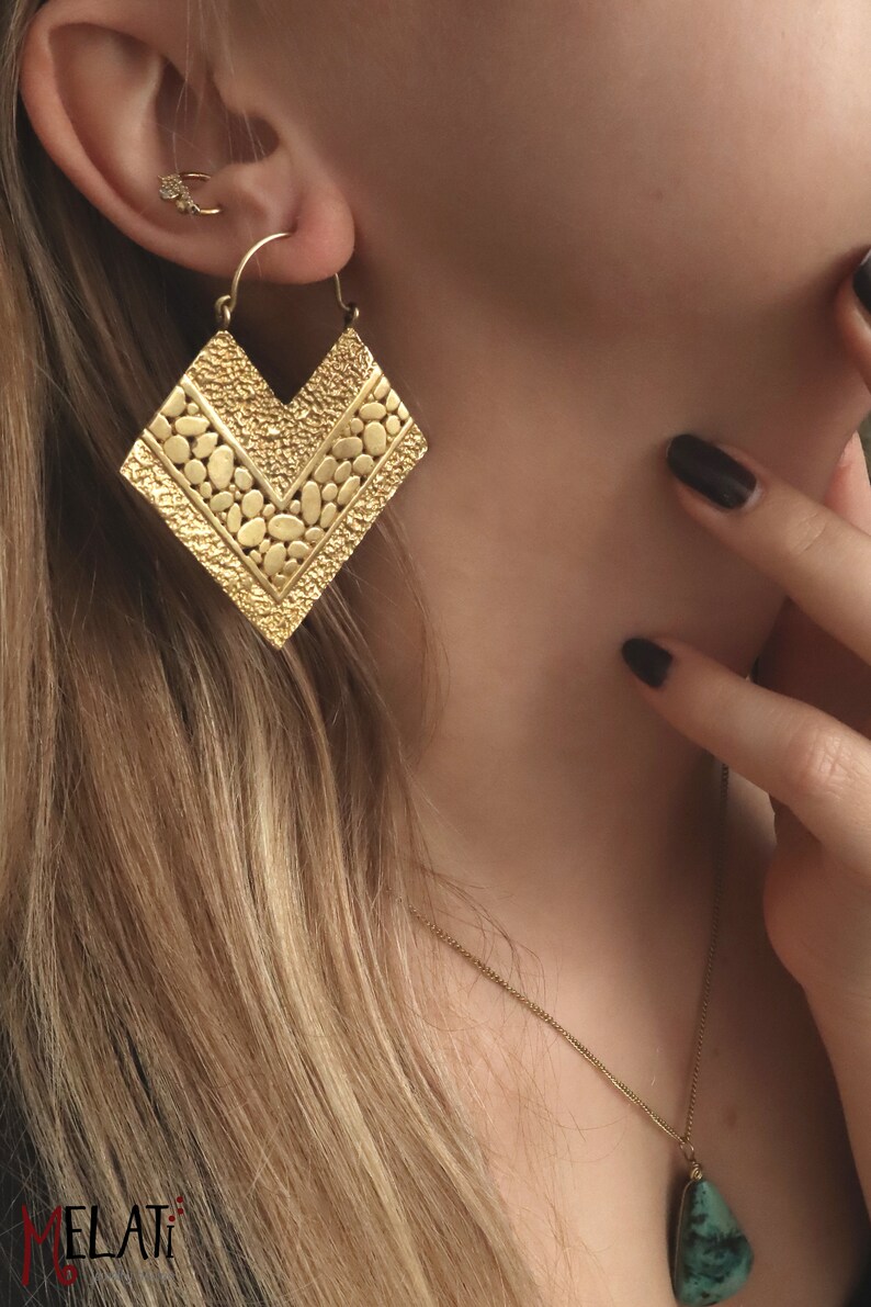May include: Gold statement earrings with a geometric, textured design. The earrings are shaped like a chevron and have a hammered finish.